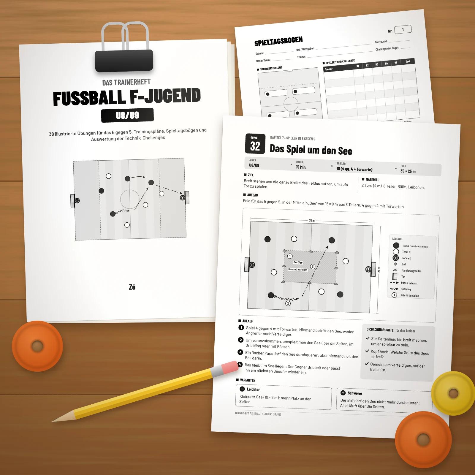 Ausgedruckte Seiten des Trainerhefts auf einem Holztisch: Titelseite mit Klammer, Spieltagsbogen und Übung Nr. 32 „Das Spiel um den See“, mit Bleistift und Markierungstellern