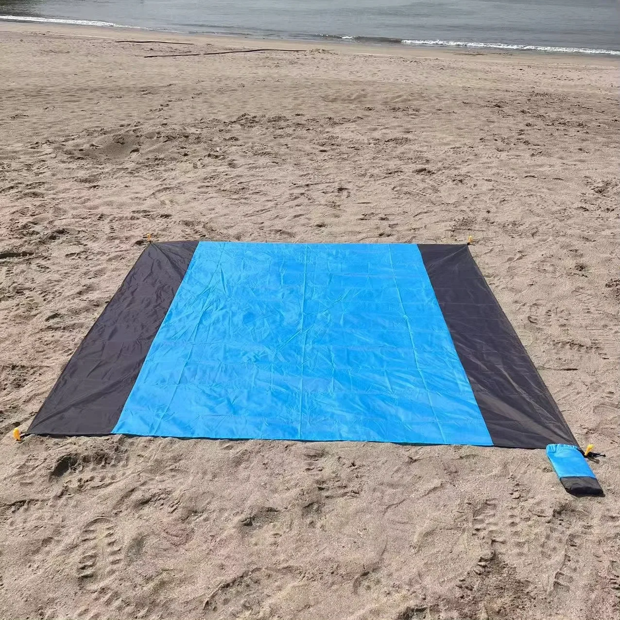 Couverture de plage bleue et grise étendue sur le sable, avec sa pochette posée dans un coin